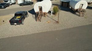 A black pickup truck is parked next to a white, covered wagon-style cabin with wooden steps, set on gravel beside a paved road. Another similar cabin is nearby.
