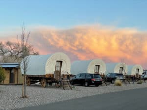 Four covered wagon-style cabins are lined up in a row on gravel, with parked cars beside them and a vibrant, orange sky in the background.