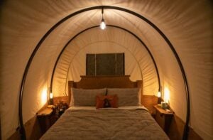 A cozy bed with neutral bedding sits inside a tent-like structure, softly lit by lamps on wooden bedside tables and a light hanging overhead.