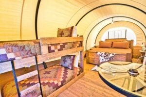 Interior of a glamping tent with wooden bunk beds and a double bed, patchwork bedding, a small table, and wood-look flooring.