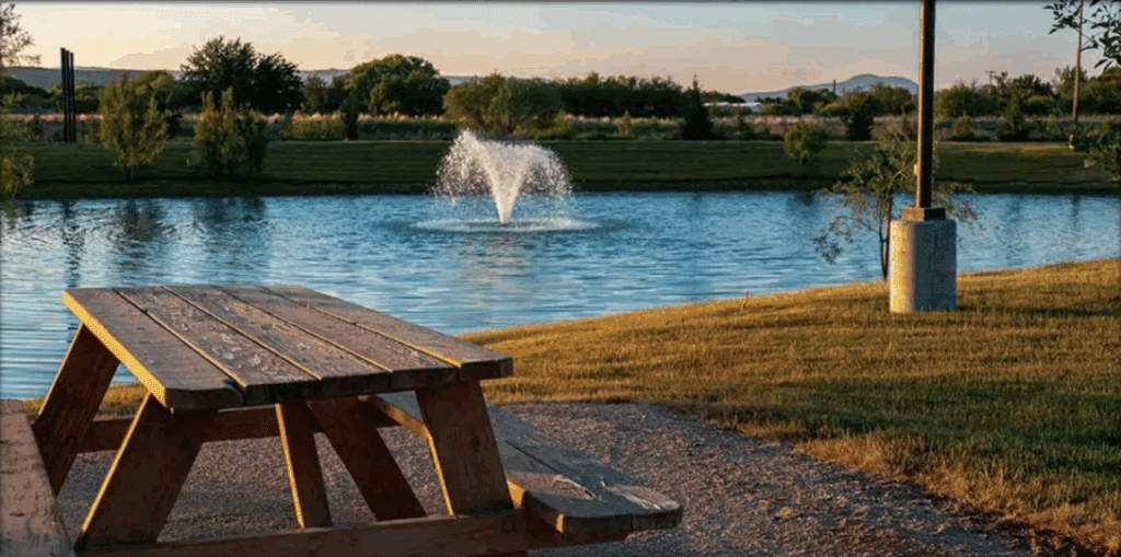 A wooden picnic table sits on a gravel area near a pond with a water fountain, surrounded by grass and trees under a clear sky.