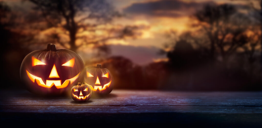 Three carved jack-o’-lanterns with glowing faces sit on a wooden surface outdoors at dusk, with trees and a colorful sunset in the background.