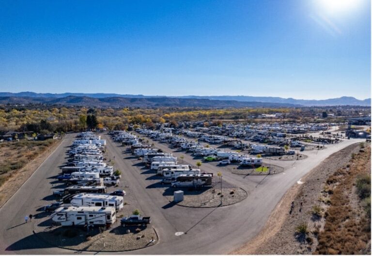 Aerial view of a large RV park with numerous recreational vehicles parked in rows under a clear blue sky, surrounded by dry, open landscape.