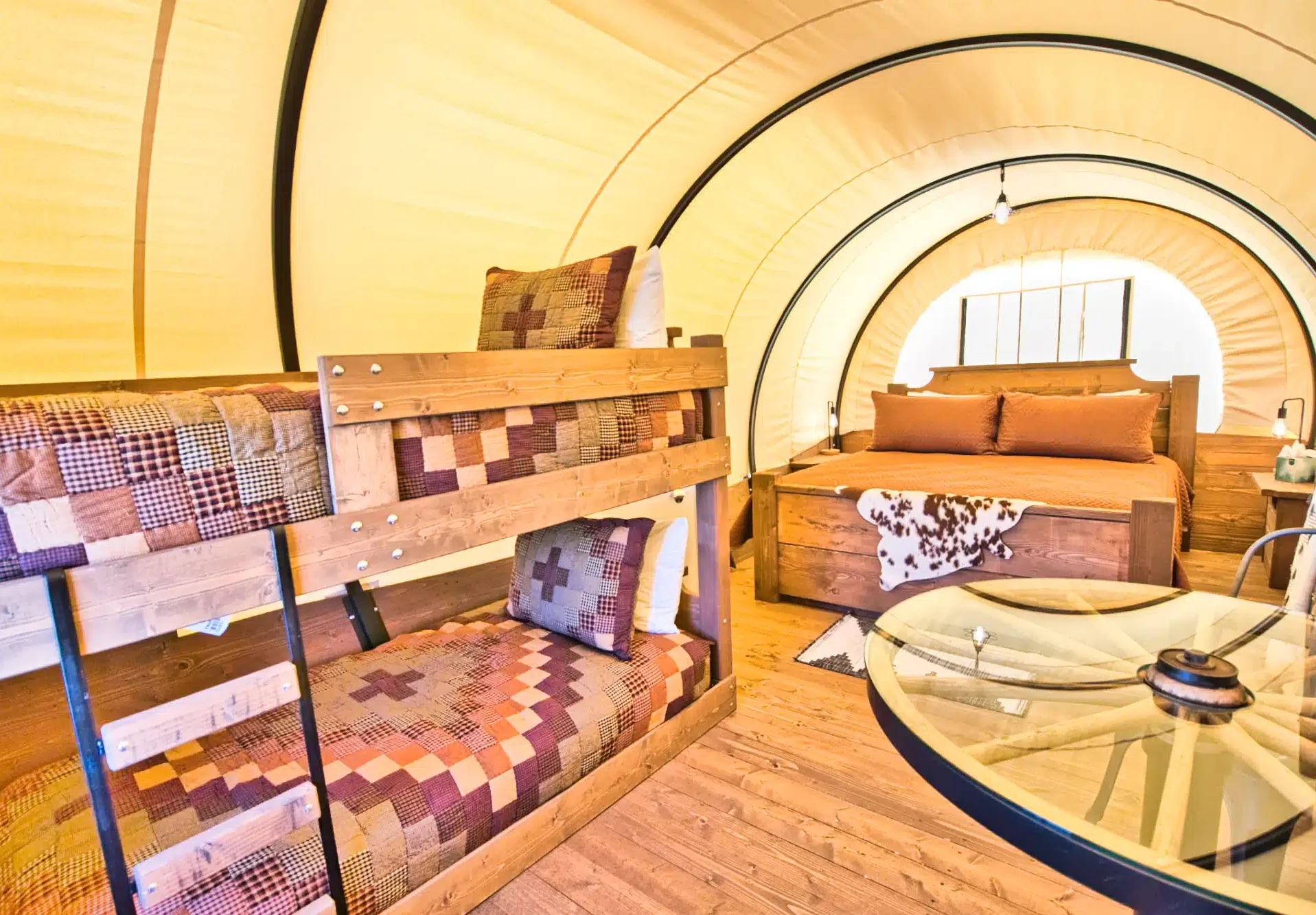 Interior of a glamping tent with wooden bunk beds, a double bed with patterned bedding, a round glass table, and wood flooring.