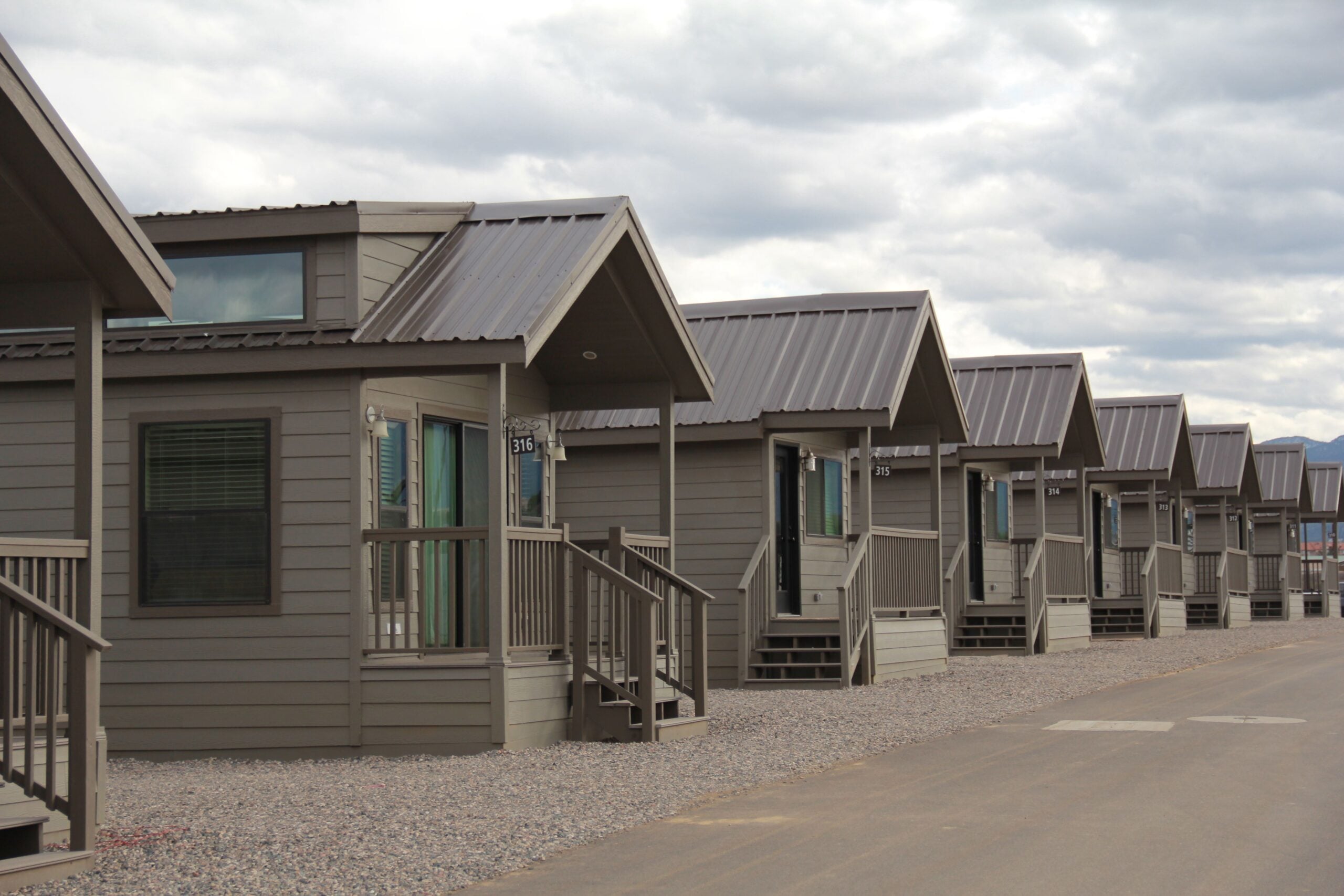 Row of identical small gray cabins with porches, set on gravel lots beside a paved road under a cloudy sky.