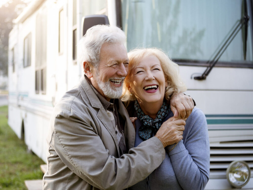 An older couple stands in front of an RV, smiling and embracing while enjoying an outdoor moment.