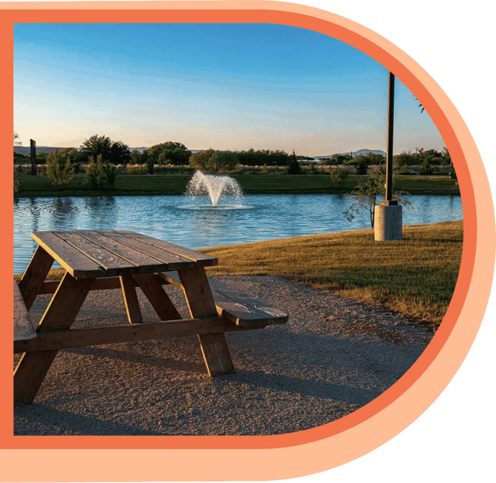 A wooden picnic table sits on a gravel area near a pond with a water fountain, surrounded by grass and trees under a clear blue sky.