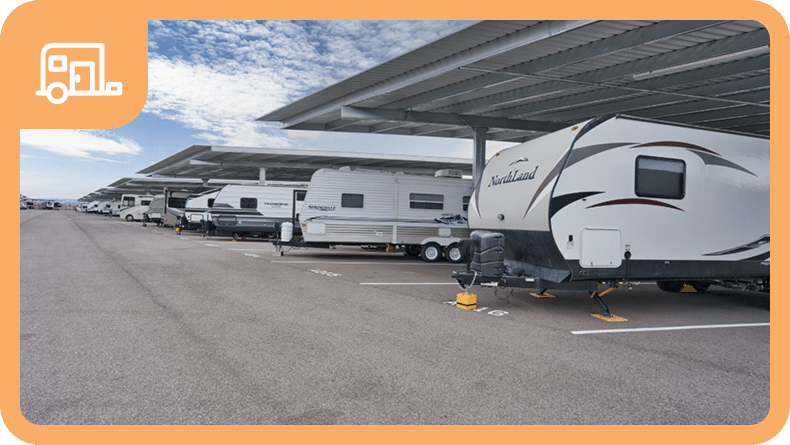 Several RVs and trailers are parked in covered parking spaces on a paved lot under a partly cloudy sky.