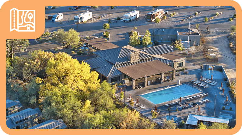 Aerial view of an RV park with several trailers, a central building, a swimming pool, poolside seating, and surrounding trees.