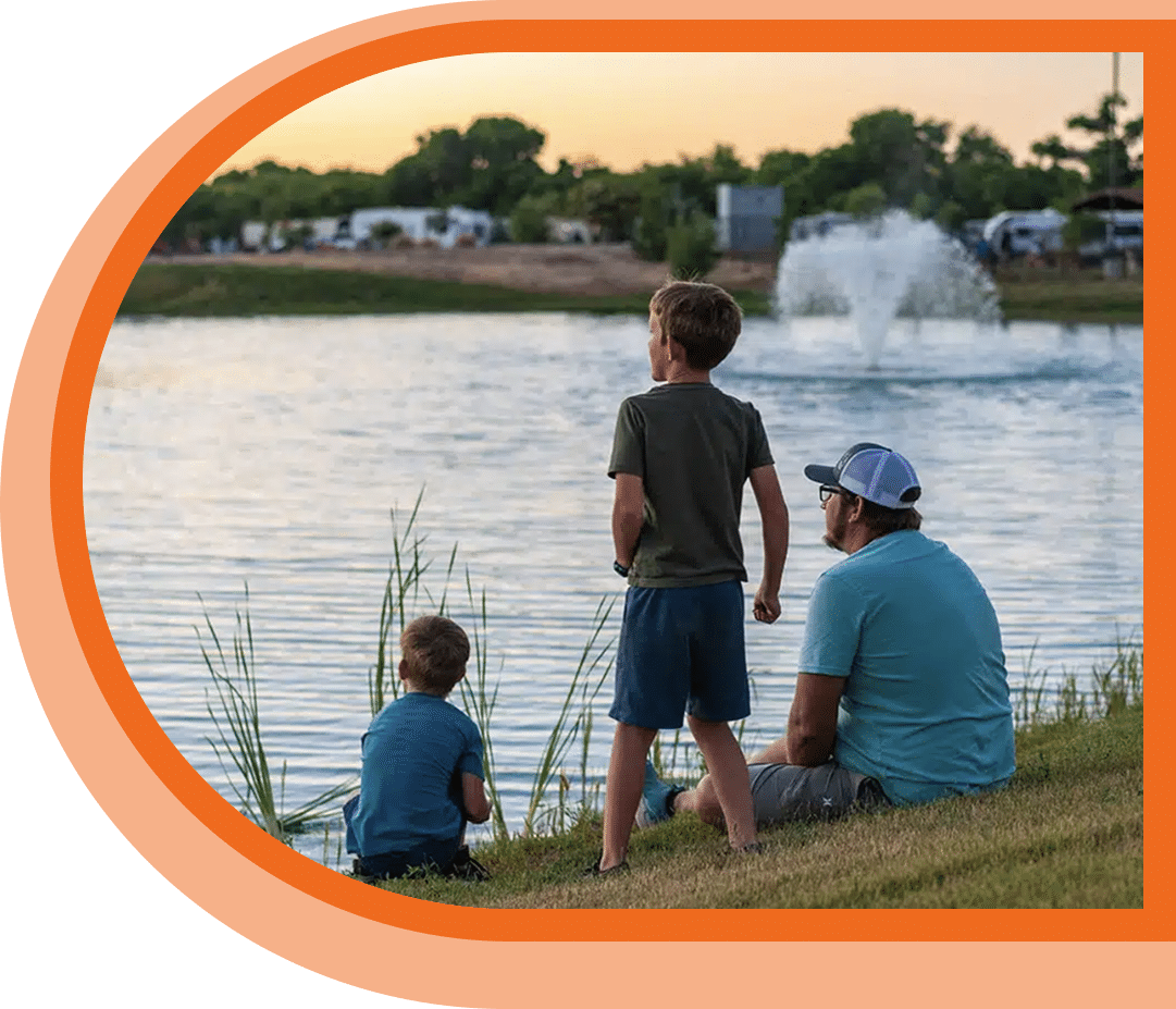 An adult and two children sit and stand by a lake, looking at a water fountain in the distance during sunset.