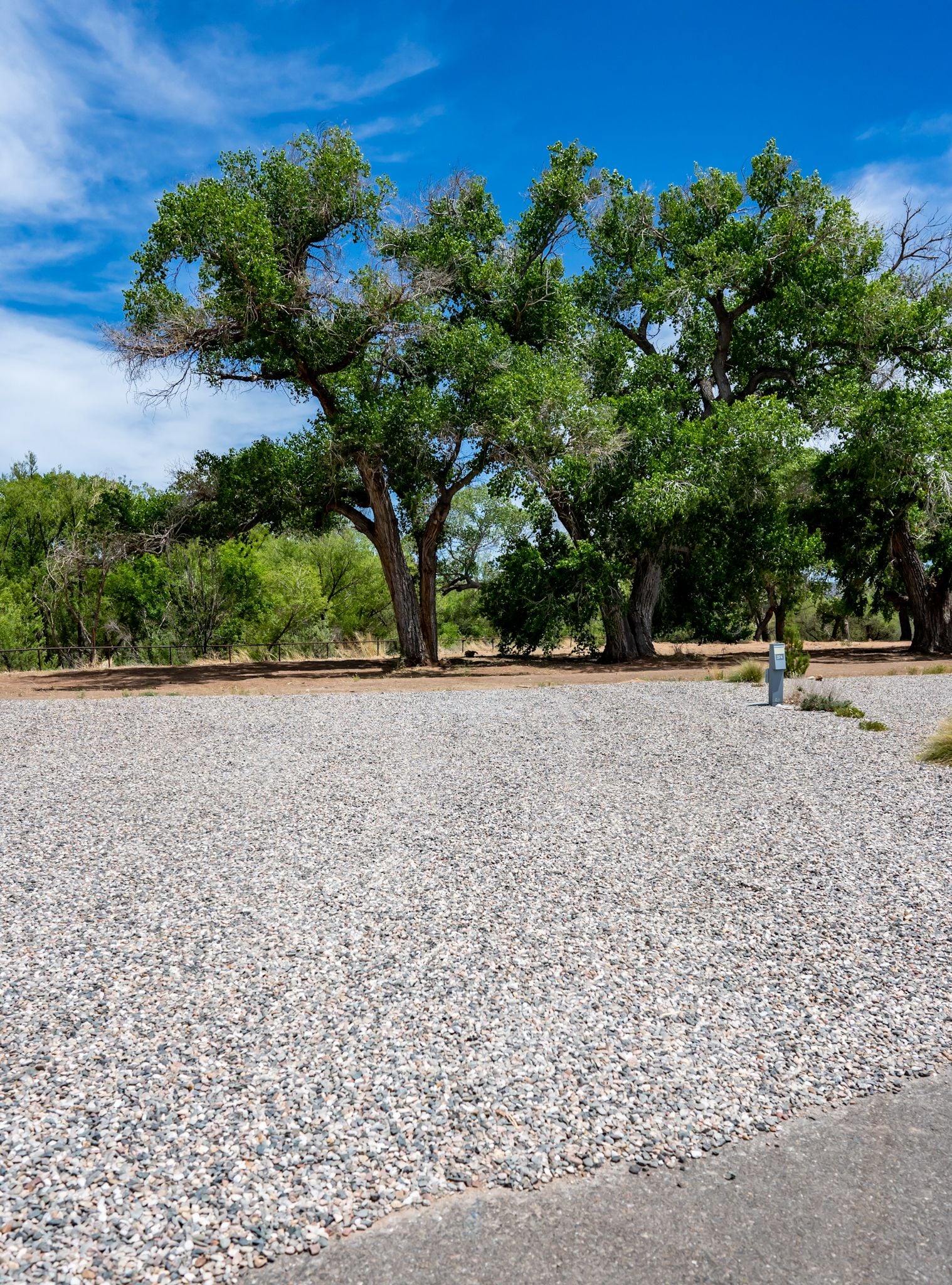 A gravel campsite with a utility hookup is surrounded by green trees under a blue sky.
