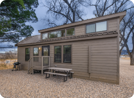 A small, modern tiny house with large windows, a front porch, and an outdoor picnic table, set on a gravel lot with bare trees and cloudy sky in the background.