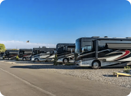 A row of large, black and silver RVs parked on gravel lots under a clear blue sky.
