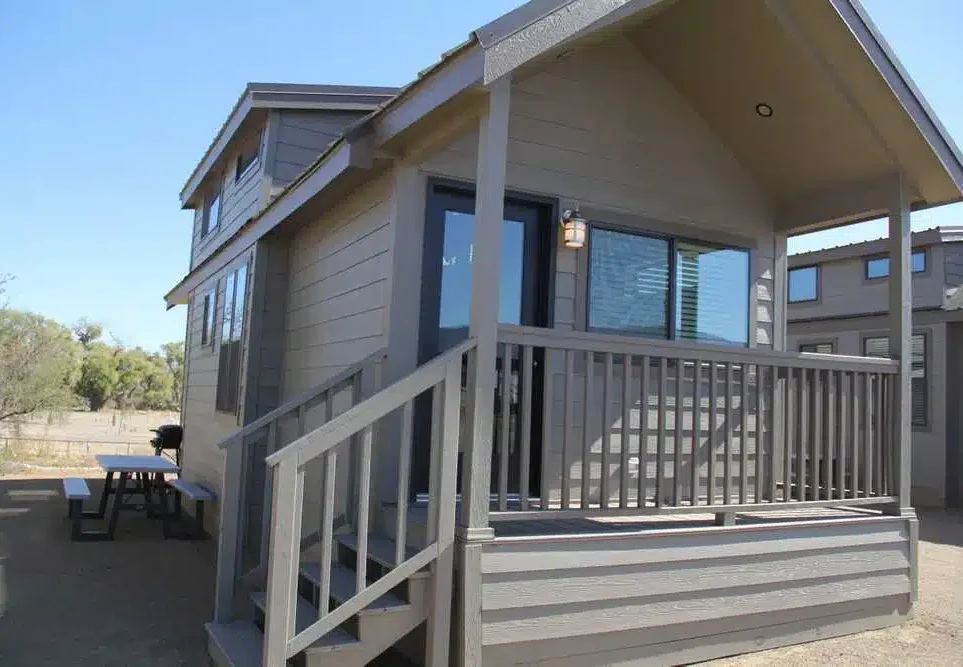 A small gray tiny house with a covered porch, front steps, and railing, situated outdoors with a picnic table and trees in the background.