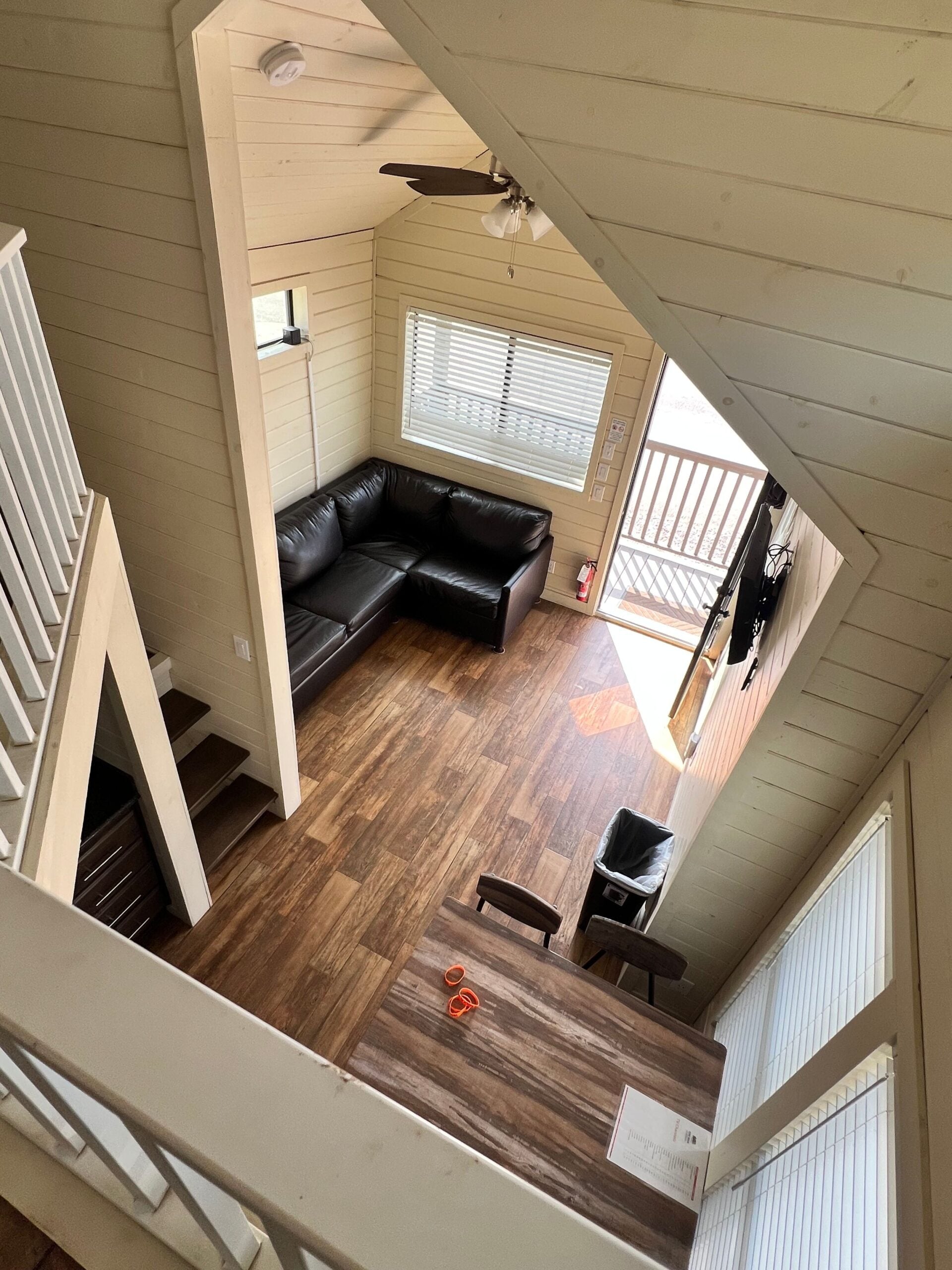 A lofted view of a small living space with wooden floors, a black sectional sofa, a dining table with two chairs, and stairs leading to a second level.