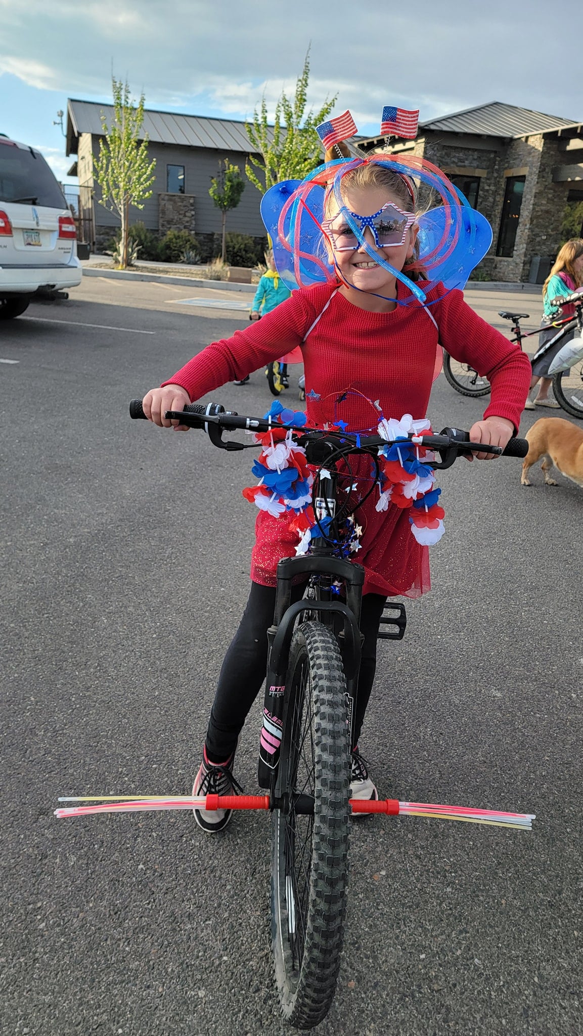 A child wearing a red dress and patriotic decorations rides a bicycle adorned with red, white, and blue garlands and a stick attached to the front wheel in a parking lot.