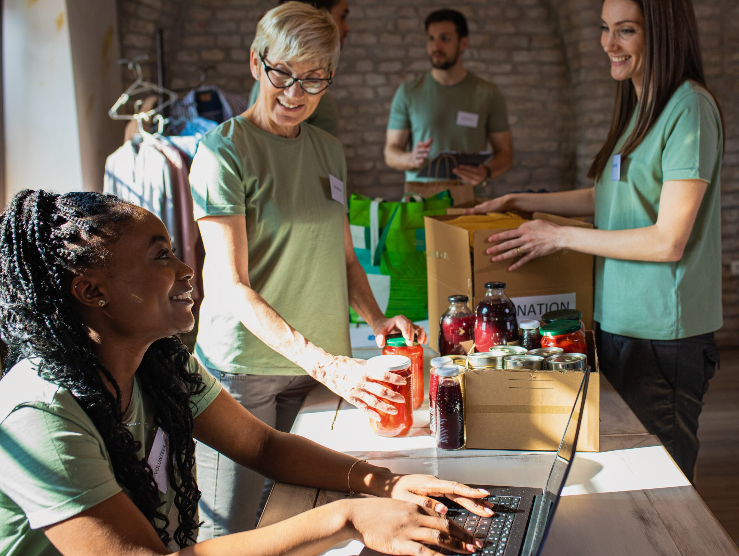 Four people in a brightly lit room pack food donations into boxes and jars, while one person works on a laptop at a table.