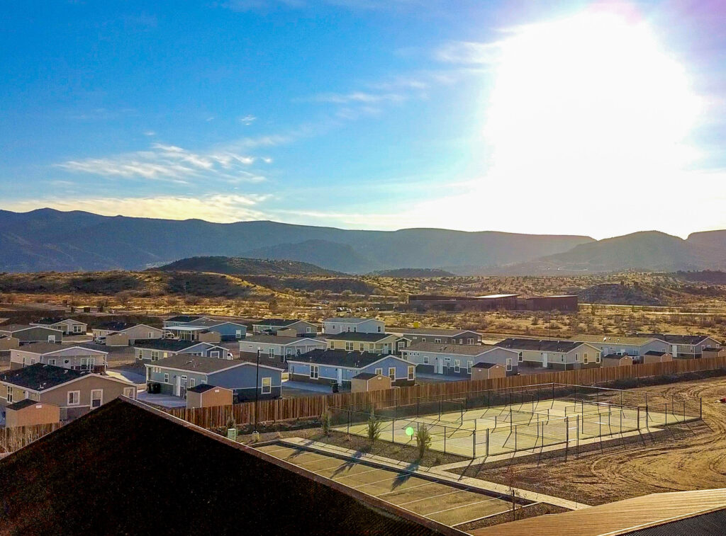 A residential neighborhood with single-story houses, a fenced tennis or basketball court, and mountains in the background under a bright, sunny sky.