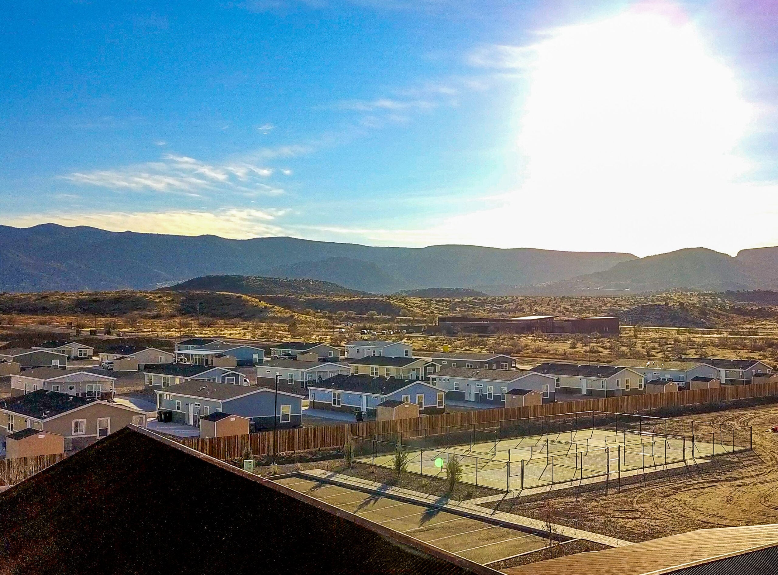A residential neighborhood with single-story houses, a fenced tennis or basketball court, and mountains in the background under a bright, sunny sky.